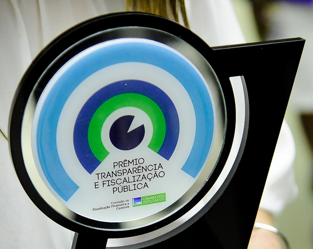 Imagem da placa comemorativa do Prêmio Transparência e Fiscalização Pública 2014, com círculos concêntricos azul-claro, branco, azul-escuro e verde.