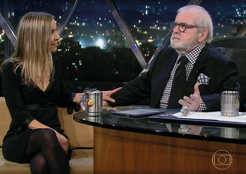 Captura de imagem do programa Jô Soares Onze e Meia: Dra Lúcia Willadino Braga, de vestido preto, conversa com o apresentador Jô Soares, vestindo camisa xadrez preta e branca com terno e gravata pretos.