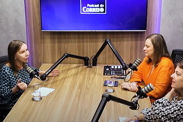 À esquerda e sentada, Dra. Lúcia Willadino Braga, vestindo blusa xadrez preta e branca, fala ao microfone. À direita, as jornalistas Denise Rothenburg, de blusa laranja, e Ana Maria Campos, de blusa com detalhes pretos e brancos, escutam.