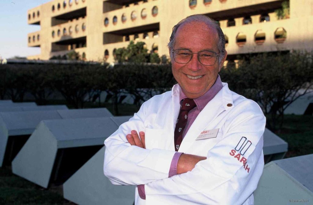 Vestindo jaleco branco, camisa lilás e gravata vinho, Dr. Aloysio Campos da Paz posa, sorridente e de braços cruzados, em frente ao SARAH Brasília Centro.