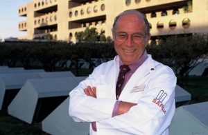 Vestindo jaleco branco, camisa lilás e gravata vinho, Dr. Aloysio Campos da Paz posa, sorridente e de braços cruzados, em frente ao SARAH Brasília Centro.