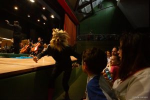 Em primeiro plano, no palco iluminado do Teatro SARAH, integrante do Ballet de Brasília dança em performance da fábula musical “Pedro e o Lobo”. Ao fundo, membros da Orquestra Sinfônica do Teatro Nacional Cláudio Santoro tocam seus instrumentos.