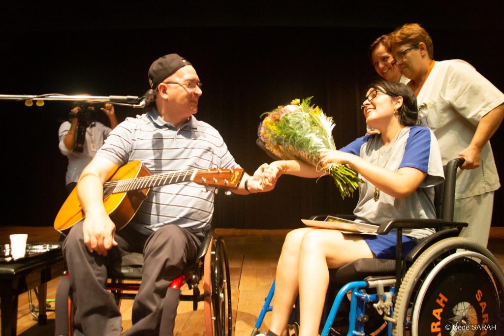 Herbert Vianna recebendo flores de uma paciente do SARAH Brasília À esquerda, sentado em cadeira de rodas, no palco iluminado do Teatro SARAH, trajando camisa polo listrada azul, calça jeans preta e boné preto com a aba voltada para trás, e segurando um violão embaixo do braço direito, Herbert Vianna aperta a mão de jovem paciente, que lhe entrega um buquê de flores. Ela, trajando uniforme hospitalar e também sentada em cadeira de rodas, sorri para o artista. À direita e mais atrás, acompanhante e colaboradora, ambas trajando uniformes, acompanham a cena e sorriem.