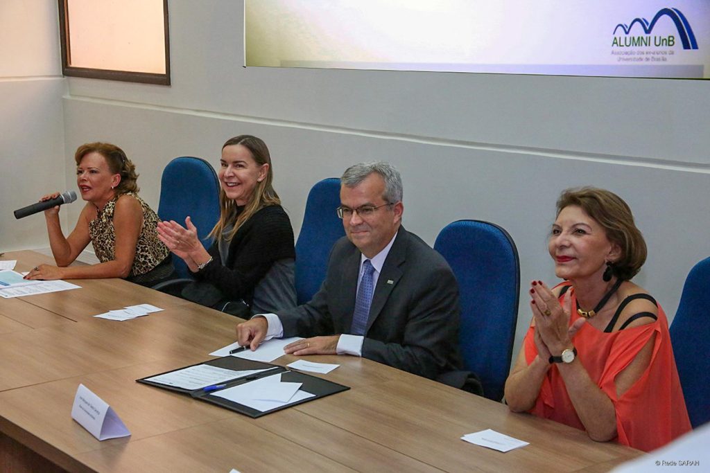Imagem de reunião especial da Alumni UnB – Associação dos Ex-alunos da Universidade de Brasília, mostra homens e mulheres sentados à uma mesa e frente a um auditório.