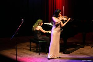 De pé, trajando um vestido longo de cor creme, a violinista Aisha Syed Castro apresenta-se no palco do Teatro SARAH, acompanhada pela pianista Lígia Monteiro, que, sentada, toca um piano de cauda.