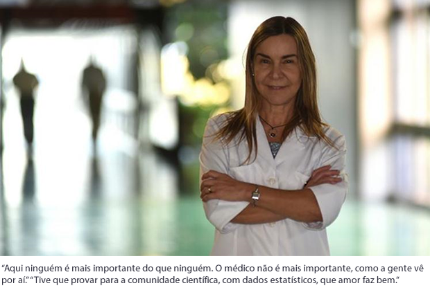 De pé, vestindo jaleco branco e de braços cruzados, Lúcia Willadino Braga posa para foto em local não identificado, com o fundo da imagem desfocado.