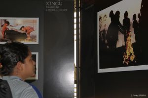 Trajando uniformes hospitalares, pacientes e acompanhantes do SARAH Belo Horizonte, alguns em cadeiras de rodas, observam um conjunto de fotos de indígenas brasileiros, dispostas em painéis, com os dizeres "Xingu –Tradição e Modernidade".