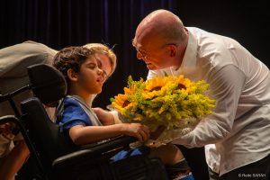 Sorrindo e trajando camisa social branca e calças pretas, o pianista Luiz Blumenschein recebe flores de menino em cadeira de roras, que traja uniforme de paciente hospitalar, no palco iluminado do Teatro SARAH Brasília.