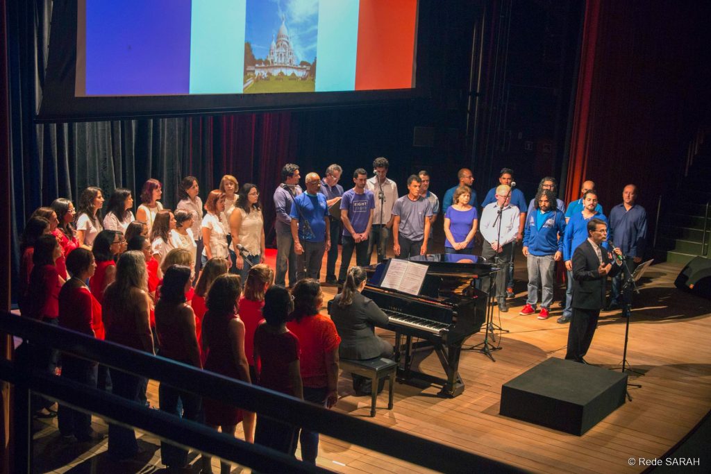 Maestro Carlos Ilha falando à plateia do Teatro SARAH No centro do palco, de pé e trajando terno escuro e gravata, o maestro Carlos Ilha fala à plateia do Teatro SARAH. Mais ao fundo, pianista sorri enquanto observa o maestro, sentada à frente de um piano de cauda. Ao seu redor, em um semicírculo, integrantes do Coral TST/Capes, todos de pé e trajando roupas vermelhas, azuis e brancas, em homenagem às cores da França, ouvem a fala do maestro atentamente.