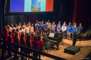 No centro do palco, de pé e trajando terno escuro e gravata, o maestro Carlos Ilha fala à plateia do Teatro SARAH. Mais ao fundo, pianista sorri enquanto observa o maestro, sentada à frente de um piano de cauda. Ao seu redor, em um semicírculo, integrantes do Coral TST/Capes, todos de pé e trajando roupas vermelhas, azuis e brancas, em homenagem às cores da França, ouvem a fala do maestro atentamente.