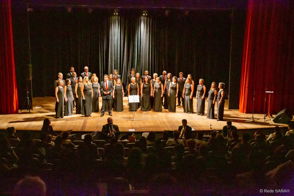Coral do Senado performando Trajando ternos pretos com camisas brancas e gravatas vermelhas ou vestidos longos pretos, membros do Coral do Senado apresentam-se, de pé e dispostos em semicírculo no palco do Teatro SARAH.
