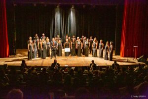 Trajando ternos pretos com camisas brancas e gravatas vermelhas ou vestidos longos pretos, membros do Coral do Senado apresentam-se, de pé e dispostos em semicírculo no palco do Teatro SARAH.