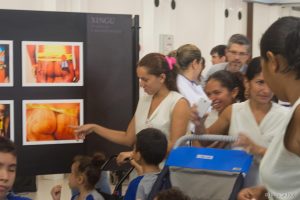 Vestindo uniforme da Rede SARAH, acompanhante aponta para a foto das nádegas de um indígena, exposta em um mural com os dizeres "Xingu", enquanto fala a crianças, também uniformizadas.
