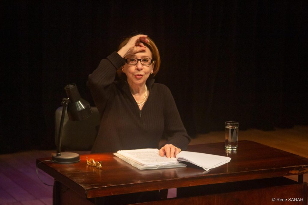 Fernanda Montenegro fazendo a leitura de “Nelson Rodrigues Por Ele Mesmo” Sentada a uma mesa de madeira escura e trajando um pulôver preto, no centro do palco iluminado do Teatro SARAH, Fernanda Montenegro faz uma leitura dramática de textos de Nelson Rodrigues.