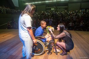 Após apresentação no Teatro SARAH Brasília, intérprete Marina Elali, trajando vestido curto preto, com brilhos, e mangas transparentes, agacha-se no palco para receber flores de paciente cadeirante. Ao fundo, pacientes, acompanhantes e colaboradores aplaudem na plateia.