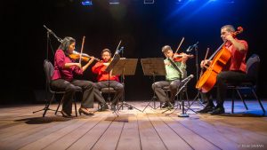 Sentados lado a lado, no palco iluminado do Teatro SARAH, apresentam-se, da esquerda para a direita, uma violinista, trajando blusa vermelha e calças pretas, dois violinistas homens, trajando camisas sociais, uma vermelha, outra verde, com calças pretas, e um violoncelista, trajando camisa vermelha de mangas curtas com calças pretas.
