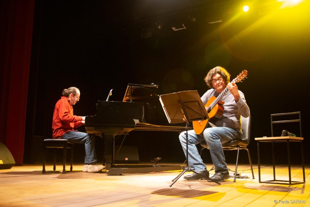 João Lucas, no piano, e Jaime Dias, no violão O pianista português João Lucas (esq.), trajando camisa social de mangas compridas vermelha e calças jeans, e o violonista brasileiro Jaime Ernest Dias, trajando camiseta de algodão de mangas longas cinza e calças jeans, apresentam-se no centro do palco iluminado do Teatro SARAH Brasília.