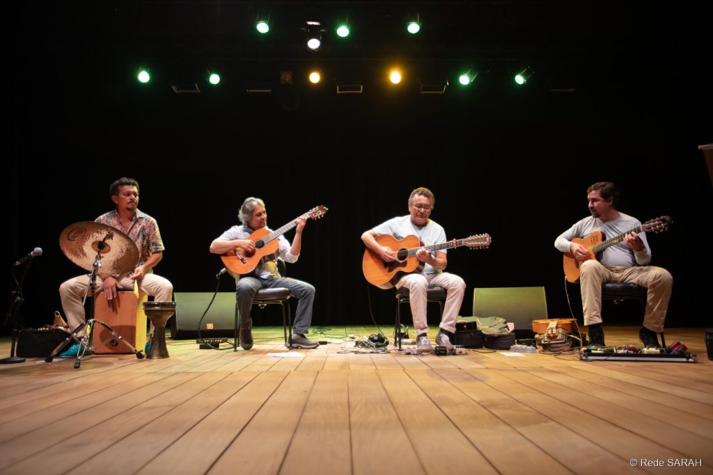 Da esquerda para direita Carlos Pial, Bosco Oliveira, Cacau Alencar e Manassés de Sousa Sentados lado a lado, no palco iluminado do Teatro SARAH apresentam-se três violonistas, trajando camisetas de algodão e calças compridas de diferentes cores e tecidos, e um percussionista, trajando camisa de mangas curtas com estampas florais e calças de tecido claro.