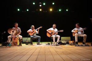 Sentados lado a lado, no palco iluminado do Teatro SARAH apresentam-se três violonistas, trajando camisetas de algodão e calças compridas de diferentes cores e tecidos, e um percussionista, trajando camisa de mangas curtas com estampas florais e calças de tecido claro.