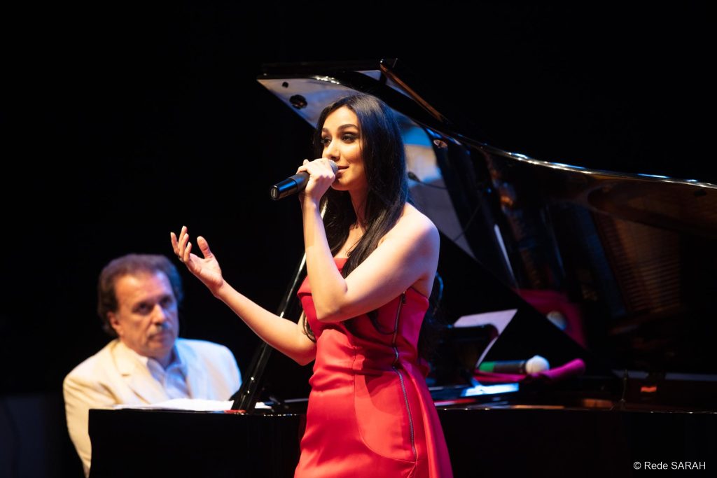 Marina Elali cantando Trajando um vestido longo de cetim vermelho, Marina Elali canta ao microfone, no palco do Teatro SARAH. Ao fundo, o maestro Eduardo Lages acompanha ao piano cauda.