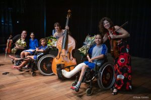Ao final da apresentação do Trio de Cordas, na coxia do Teatro SARAH Brasília, integrantes do grupo posam para fotos com pacientes cadeirantes. As musicistas têm nas mãos seus instrumentos e ramalhetes de flores, e trajam vestidos longos, com estampas coloridas, enquanto os pacientes vestem uniformes hospitalares.