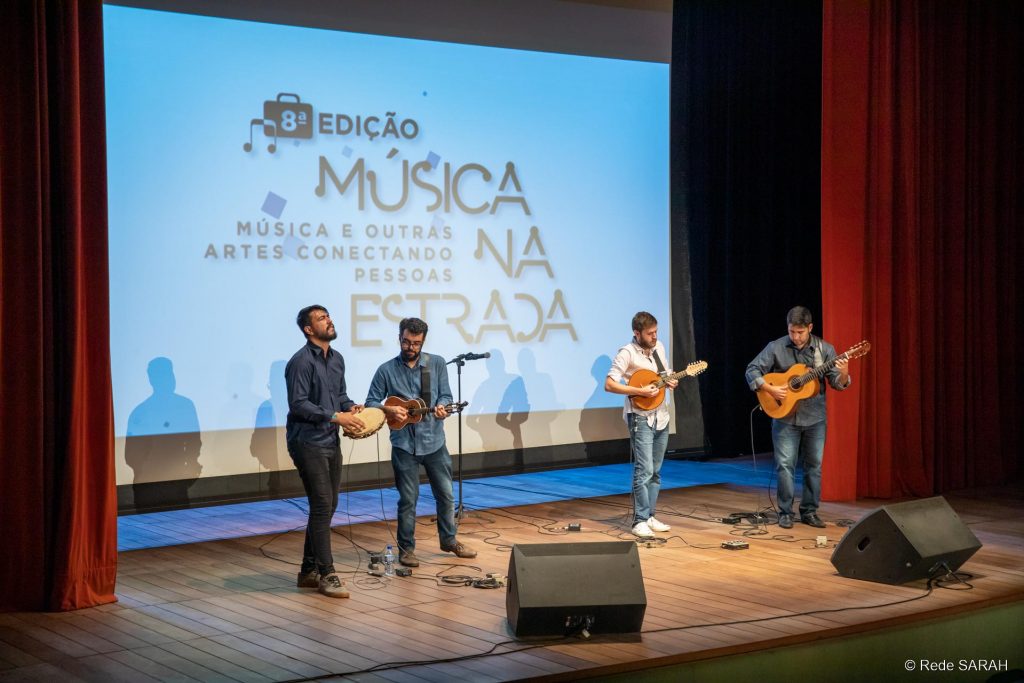 Grupo de Choro Sai da Frente performando juntos No palco iluminado do Teatro SARAH, de pé, apresentam-se quatro músicos, tocando pandeiro, cavaquinho, bandolim e violão. Trajam calças jeans, camisas de mangas compridas e tênis. Ao fundo, grande tela de projeção mostra os dizeres "8ª Edição – Música na Estrada, Música e Outras Artes Conectando as Pessoas”.