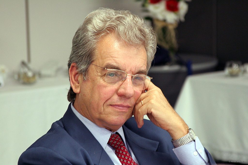 Carlos Átila aparece sentado, vestindo terno azul-marinho e gravata vermelha, tocando o próprio rosto com a mão esquerda, em uma sala de reuniões.