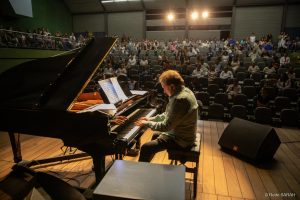 Em primeiro plano, no palco iluminado do Teatro SARAH, o maestro Eduardo Lages, sentado, toca um piano de cauda no Teatro SARAH Brasília. Lages traja uma camisa social de linho verde-clara e calças jeans pretas. Ao fundo, na penumbra, aparece o auditório e a plateia de pacientes, acompanhantes e colaboradores da Rede SARAH.