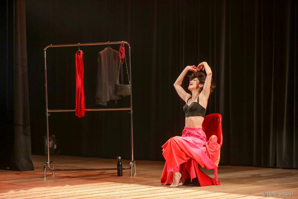 Juliana Martins apresentando a peça “O prazer é todo nosso” No centro do palco, sentada em uma cadeira vermelha, descalça e vestindo um top preto com uma saia vermelha longa e bufante, a atriz Juliana Martins sorri e levanta os braços, lançando ao ar os cabelos.