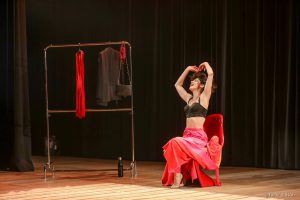 No centro do palco, sentada em uma cadeira vermelha, descalça e vestindo um top preto com uma saia vermelha longa e bufante, a atriz Juliana Martins sorri e levanta os braços, lançando ao ar os cabelos.