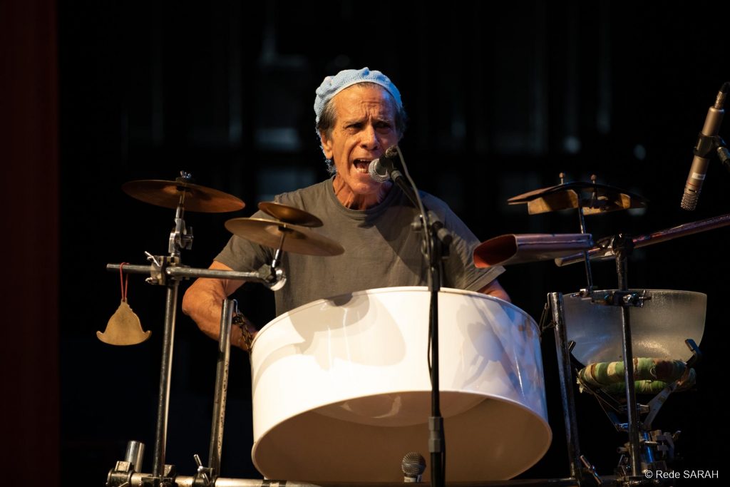 Sérgio Boré performando sua apresentação Close mostra o percussionista Sérgio Boré cantando ao microfone e tocando um tambor de aço. Iluminado, no palco do Teatro SARAH, ele traja camiseta cinza e boina azul-clara.
