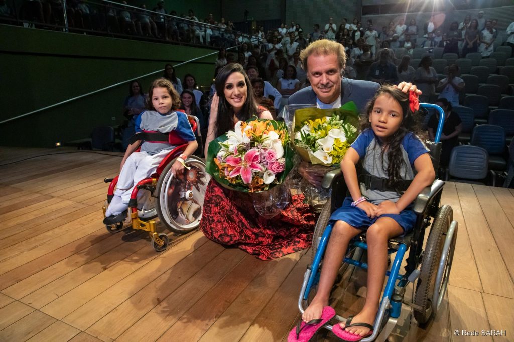 Eduardo Lages e Marina Elali posando para foto junto às pacientes do SARAH Brasília e o público do Teatro SARAH Logo após apresentação no Teatro SARAH, Eduardo Lages e Marina Elali, agachados e segurando ramalhetes de flores, posam com crianças pacientes do SARAH Brasília, ambas em cadeiras de rodas, no palco.