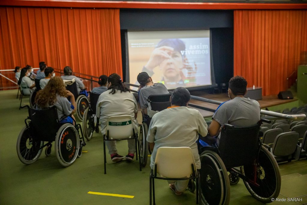 Público do Teatro SARAH Brasília assistindo à palestra Em primeiro plano, em uma das galerias laterais do Teatro SARAH, pacientes em cadeiras de rodas, acompanhantes e colaboradores da Rede acompanham apresentação. Ao fundo, no palco, grande tela de projeção mostra imagem de uma mulher gesticulando em Libras e os dizeres "Assim Vivemos – 10º Festival Internacional de Filmes sobre Deficiência".