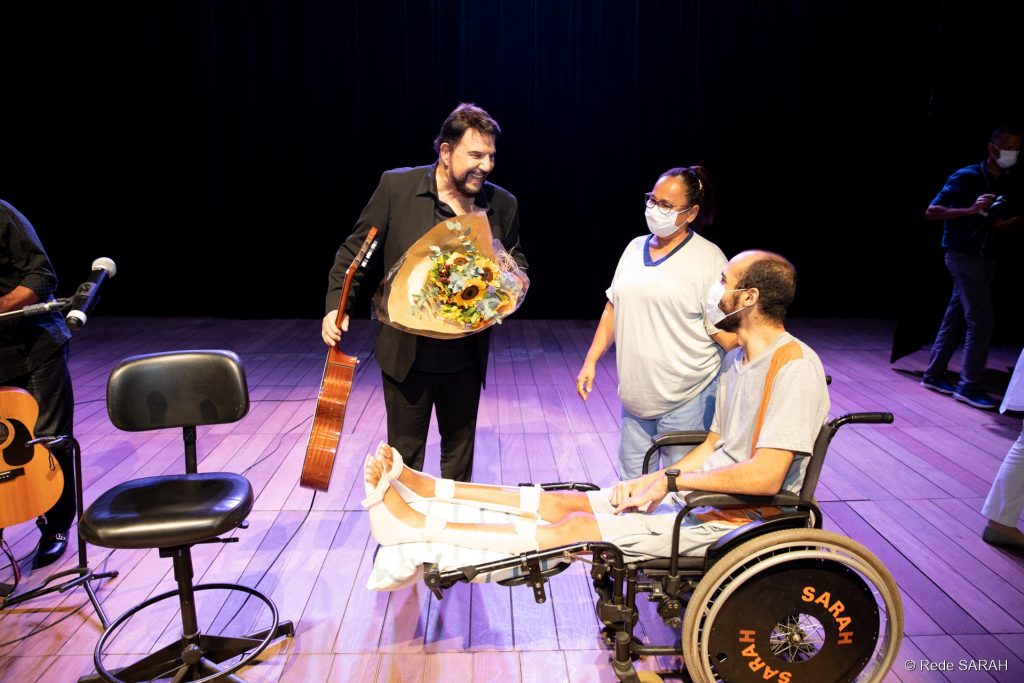 Michael Sullivan agradecendo as flores recebidas de um paciente do SARAH Brasília De pé, no palco do Teatro SARAH, trajando um terno e camisa social pretos, sem gravata, e segurando um violão na mão direita e um buquê de flores na esquerda, Michael Sullivan conversa com paciente em cadeira de rodas e sua acompanhante.