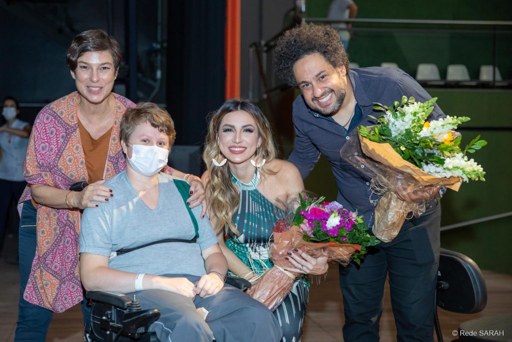 Atriz Maria Paula (à esquerda), Juliani Fernandes e Diego Figueiredo posando para foto junto à paciente do SARAH Brasília A atriz Maria Paula (à esquerda, vestindo calças jeans, uma bata estampada laranja e rosa, e uma blusa marrom), uma paciente do SARAH Brasília (sentada em cadeira de rodas, vestindo uniforme hospitalar), a cantora Juliani Fernandes (agachada, segurando um buquê de flores e trajando um vestido longo azul, verde e branco) e o violonista Diego Figueiredo (vestindo camisa de mangas longas azul e calças jeans) posam para foto no Teatro SARAH.