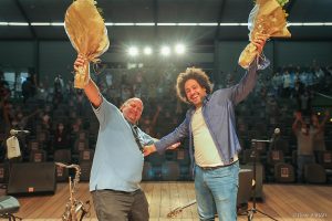 De pé, sorrindo com os braços abertos e segurando buquês de flores, Diego Figueiredo e Derico posam no palco iluminado do Teatro SARAH em Brasília. Ao fundo, a plateia de colaboradores, pacientes e acompanhantes da Rede SARAH também ergue os braços e acena para a foto. Derico traja camisa polo azul-claro e calça de brim marrom; Diego veste calça jeans, camiseta branca e camisa social azul desabotoada.