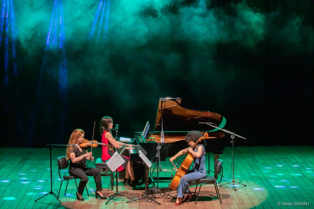 Atlas Trio performando juntas Envoltas em fumaça de gelo seco e luzes azuis e verdes, pianista, violinista e violoncelista apresentam-se no palco do Teatro SARAH em Brasília.