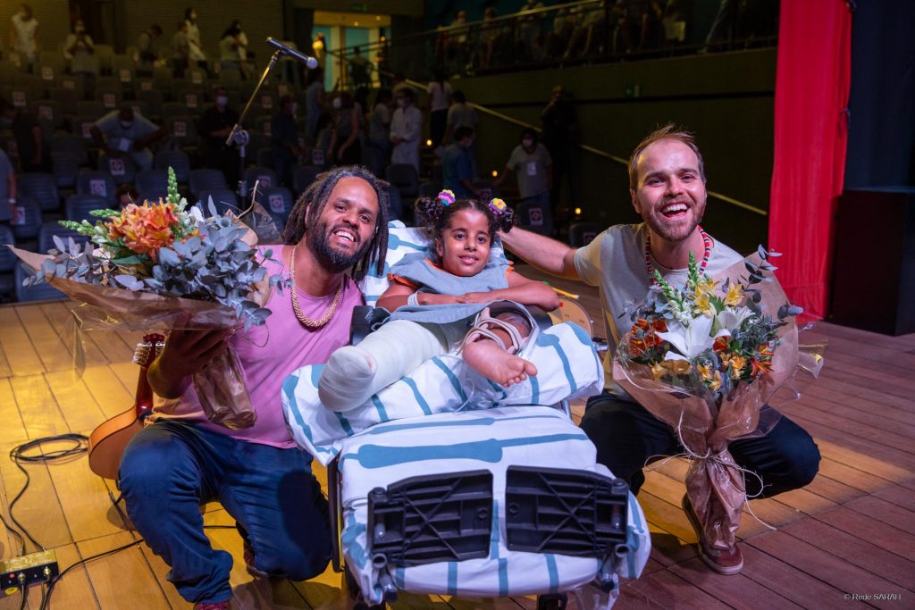 Stênio Neves e Filipe Braga posando para foto junto à paciente do SARAH Brasília O bandolinista Filipe Braga e o violonista Stênio Neves, agachados e segurando buquês de flores, posam, no palco do Teatro SARAH, com paciente do SARAH Brasília, garota deitada em maca, com uniforme do hospital e uma das pernas engessada.