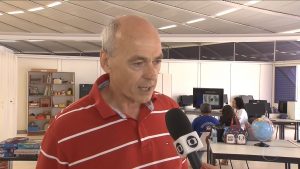 CAPTURA 20160324 Método De Tratamento Da Rede SARAH Chama Atenção De Pesquisadores Holandeses De pé e vestindo camisa polo vermelha, com listras brancas, homem fala a repórter da TV Globo no SARAH Lago Norte, em Brasília. Ao fundo, pacientes a profissionais de reabilitação conversam entre si.