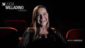 Captura de tela: de vestido preto e sentada em uma poltrona de cinema, Dra. Lúcia Willadino Braga sorri para a câmera.