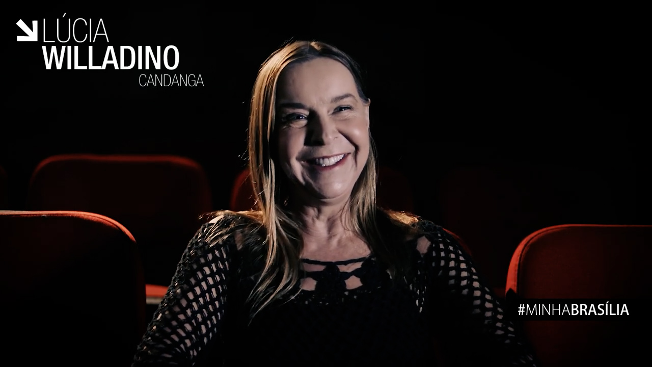 Captura de tela: de vestido preto e sentada em uma poltrona de cinema, Dra. Lúcia Willadino Braga sorri para a câmera.