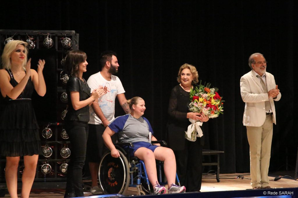 Eva Wilma agradecendo as flores recebidas de uma paciente do SARAH Brasília De pé e segurando um grande buquê de flores que acabou de receber de uma paciente sentada em cadeira de rodas, no palco do Teatro SARAH, Eva Wilma recebe palmas dos demais membros do elenco.