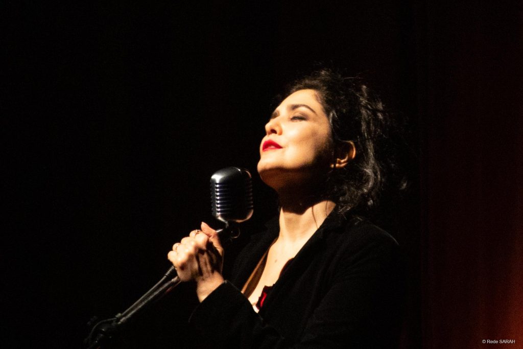 Letícia Sabatella performando Close mostra Letícia Sabatella, iluminada por um spot de luz, cercada de sombras, vestindo um sobretudo preto, cantando, de olhos fechados, enquanto segura microfone e pedestal.