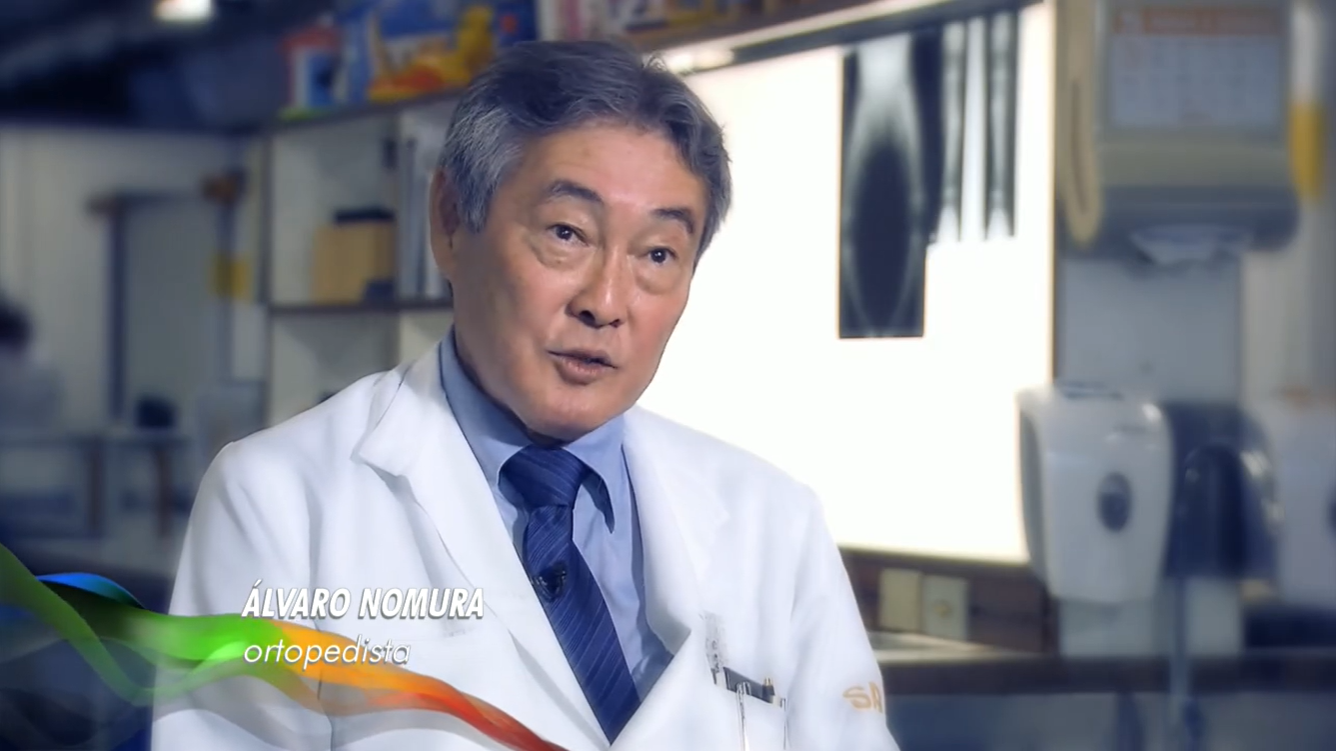 Dr. Álvaro Nomura aparece sentado e vestindo jaleco com camisa social azul e gravata azul-marinho. Ao fundo, consultório, onde aparecem radiografias de pacientes penduradas em tela luminosa.