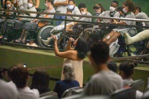 Vannick Belchior circula entre a plateia de pacientes, acompanhantes e colaboradores, enquanto canta, de pé e trajando um vestido branco longo, no Teatro SARAH.
