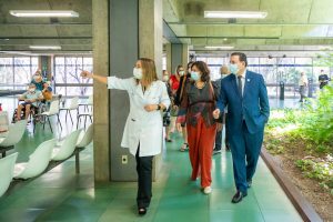 Dra. Lúcia Willadino Braga mostrando o SARAH Brasília à ministra da Saúde, Nísia Trindade Vestindo jaleco branco, Dra. Lúcia Willadino Braga apresenta o ambulatório do SARAH Brasília Centro à ministra da Saúde, Nísia Trindade, vestindo blusa de manga comprida em tons de vermelho e azul com calças vermelhas, e ao secretário-executivo da Saúde, Swedenberger Barbosa, vestindo terno azul e gravata. Ao fundo, membros da comitiva ministerial e da equipe do hospital acompanham a visita.