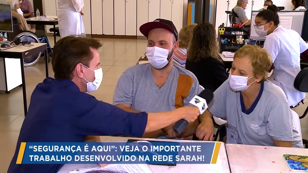 Sentado e vestindo camisa azul-marinho, repórter segura microfone para ouvir acompanhante de paciente da Rede SARAH, que veste uniforme cinza. Entre eles, paciente, com uniforme cinza e boné preto, ouve atentamente. Todos usando máscaras cirúrgicas e sentados em torno de uma mesa. Ao fundo, pacientes e profissionais de reabilitação interagem.