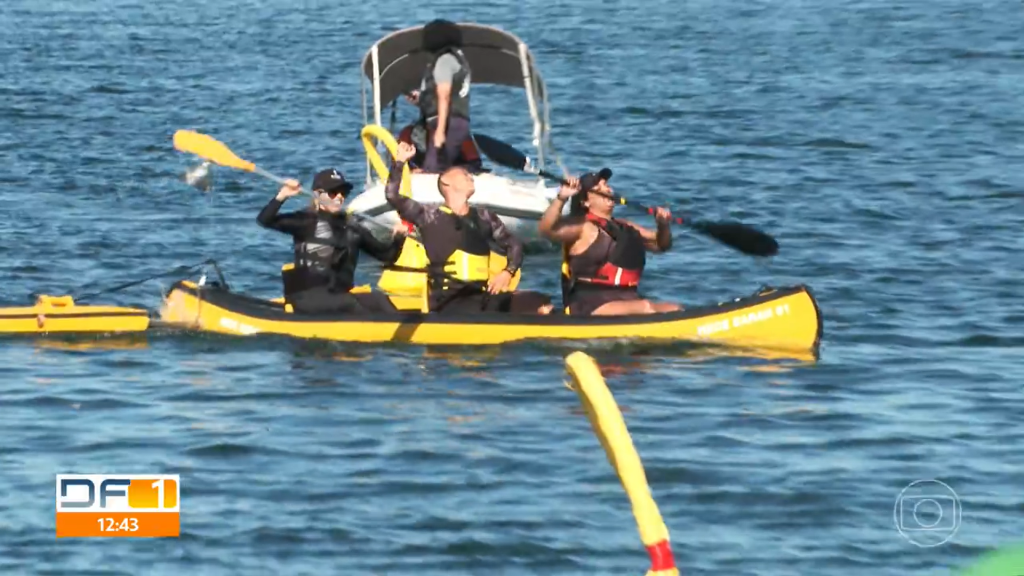 Trajando roupas pretas e coletas salva-vidas, três pacientes comemoram a conclusão de prova de paracanoagem no Lago Paranoá. Ao fundo, vê-se bote de apoio e colaboradores da Rede SARAH.