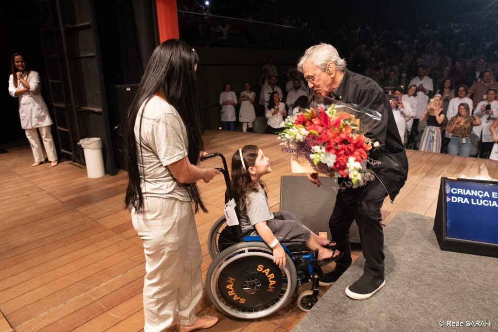 Caetano Veloso recebendo flores de Maisa Santos Bernardes, paciente do SARAH Brasília De pé e sorrindo, Caetano Veloso inclina-se para abraçar criança em cadeira de rodas, paciente do SARAH Brasília, de quem recebeu um buquê de flores.