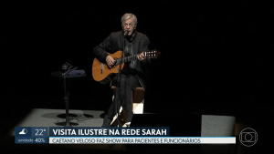 Captura de tela da TV Globo Brasília mostra Caetano Veloso sentado, trajando casaco, camisa e calças pretas, cantando ao microfone e tocando violão, no centro do palco iluminado do Teatro SARAH. Na base da imagem, vinheta mostra os dizeres: "VISITA ILUSTRE NA REDE SARAH. Caetano Veloso faz show para pacientes e funcionários."
